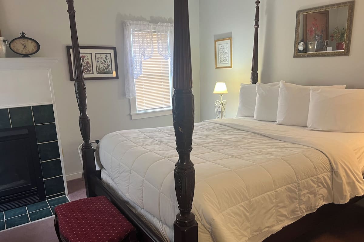 Uma cama com dossel, roupa de cama branca, um banco estofado vermelho, uma lareira e um relógio fixado na parede.