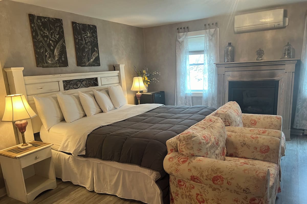 Quarto com uma cama grande, mesa de cabeceira com abajur, poltrona estampada, lareira e duas pinturas penduradas na parede.