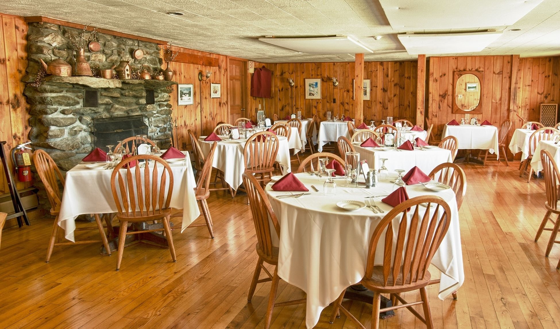 A restaurant with wooden walls, stone fireplace, and multiple tables set with white tablecloths and red napkins.