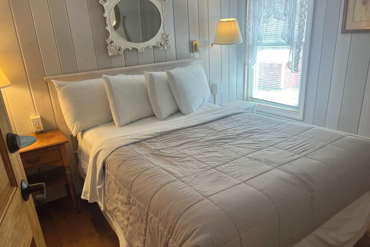Cama bem arrumada com cobertura acolchoada, mesa de cabeceira com abajur e espelho fixado na parede.