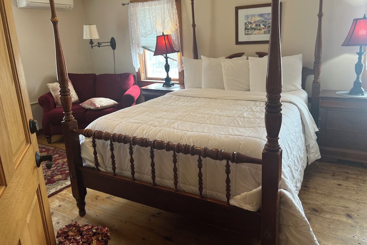 Uma cama de dossel tradicional com estrutura de madeira e roupa de cama branca, uma poltrona vermelha, uma mesa de cabeceira com abajur e uma janela com cortinas leves.