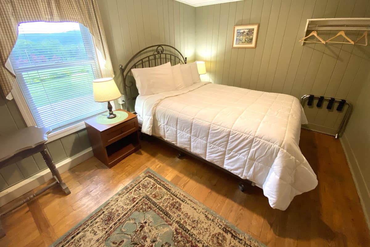 Uma cama bem arrumada em um quarto com assoalho de madeira, uma mesa de cabeceira com luminária, uma janela com persianas e um radiador.