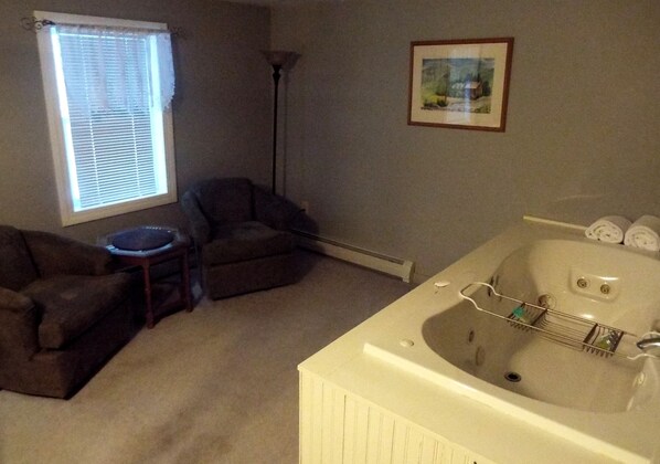 A room with a large bathtub, a window with blinds, a floor lamp, and a framed picture on the wall.