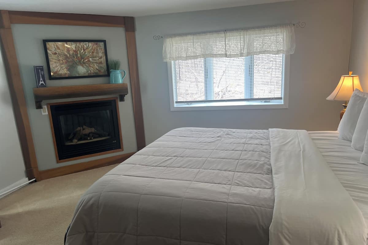 Um quarto com uma cama grande, uma lareira, uma janela com persianas e uma luminária em uma mesa de cabeceira.
