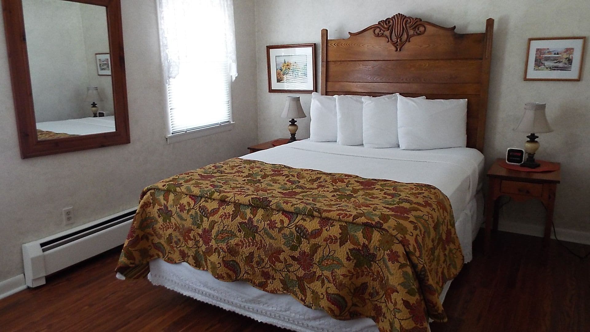 A bedroom with a wooden headboard, a floral bedspread, white bedding, a nightstand with a lamp, and a mirror on the wall.