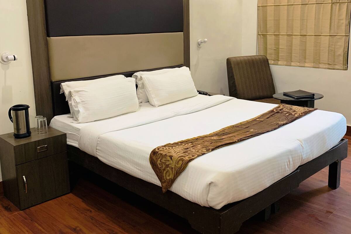 A neatly made bed with white linens and a brown decorative throw, a bedside table with a kettle and glasses, and a chair with a cushion.