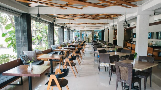 A spacious cafe with multiple seating areas, including tables and benches, and large windows offering views of the outdoors. - Bloo Bali Hotel (Legian)