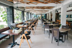 Un café amplio con varias zonas de estar, incluyendo mesas y bancos, y grandes ventanales que ofrecen vistas al exterior.