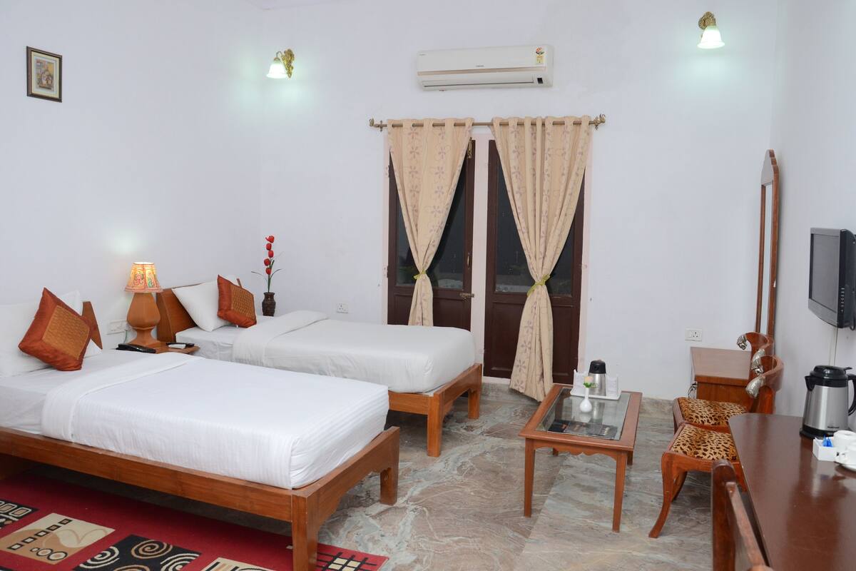 A hotel room with two beds, a desk, a chair, a TV, and an air conditioning unit.
