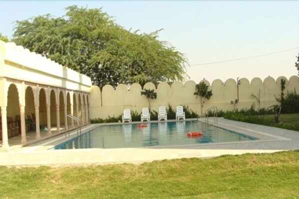 A swimming pool with a white wall and arches, surrounded by grass and trees.
