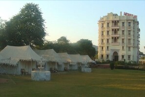 Um hotel com várias tendas brancas montadas em um pátio, um edifício grande com varandas, e uma placa indicando "Satyam Palace".