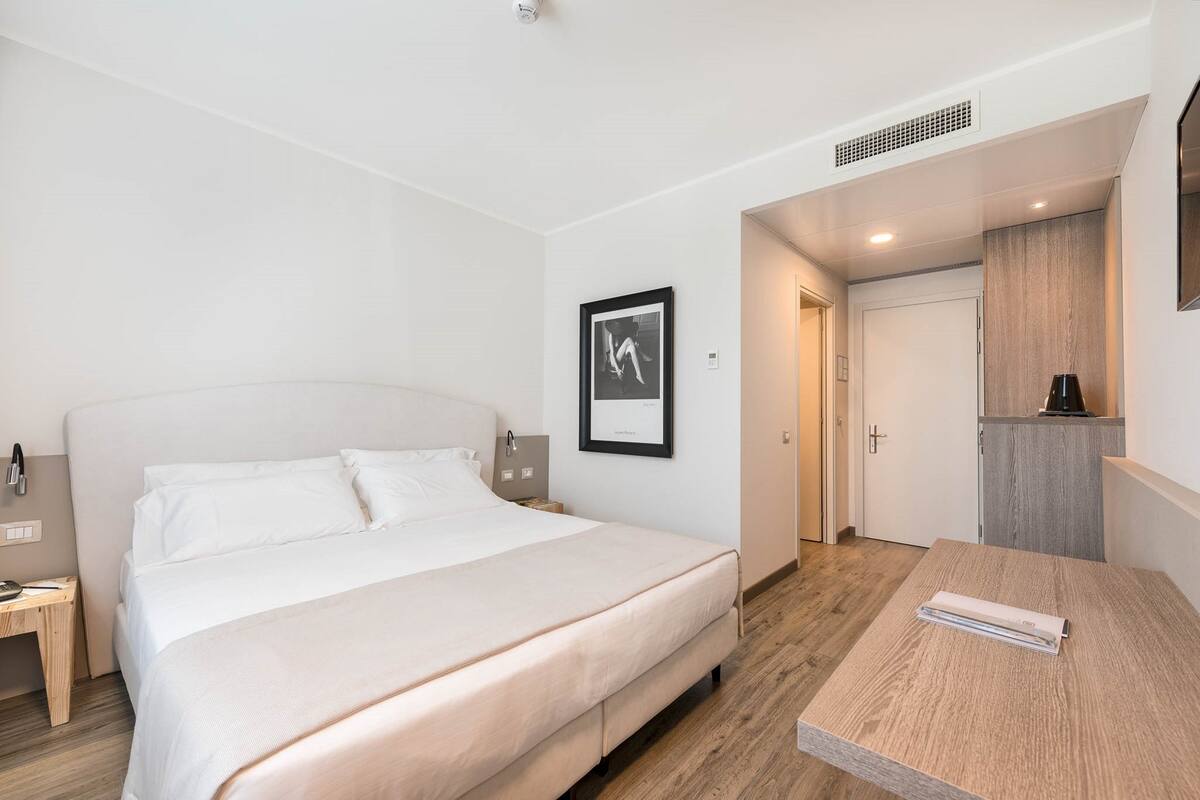 Ett modernt hotellrum med en stor säng, ett sängbord, en liten avlastningsyta, en inramad tavla på väggen och en liten köksdel.