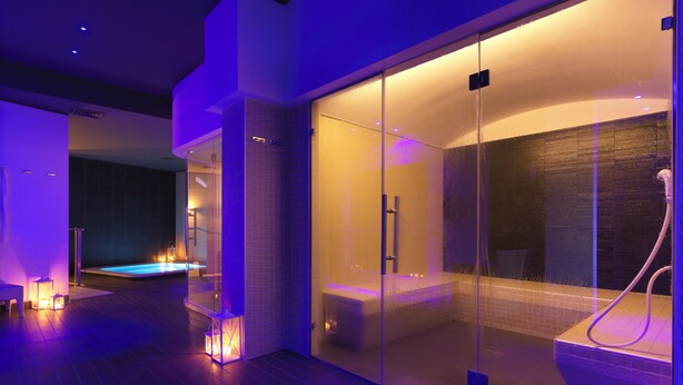 A modern spa area with a glass-enclosed hot tub, a separate shower area, and a pool visible in the background. -  DB HOTEL VERONA, AIRPORT CONGRESS & SPA (Sommacampagna)