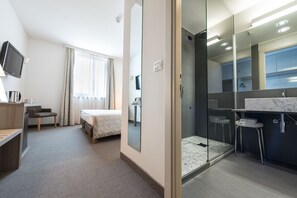 A hotel room with a bed, a desk, a TV, a shower, and a bathroom with a mirror and sink. -  DB HOTEL VERONA, AIRPORT CONGRESS & SPA (Sommacampagna)