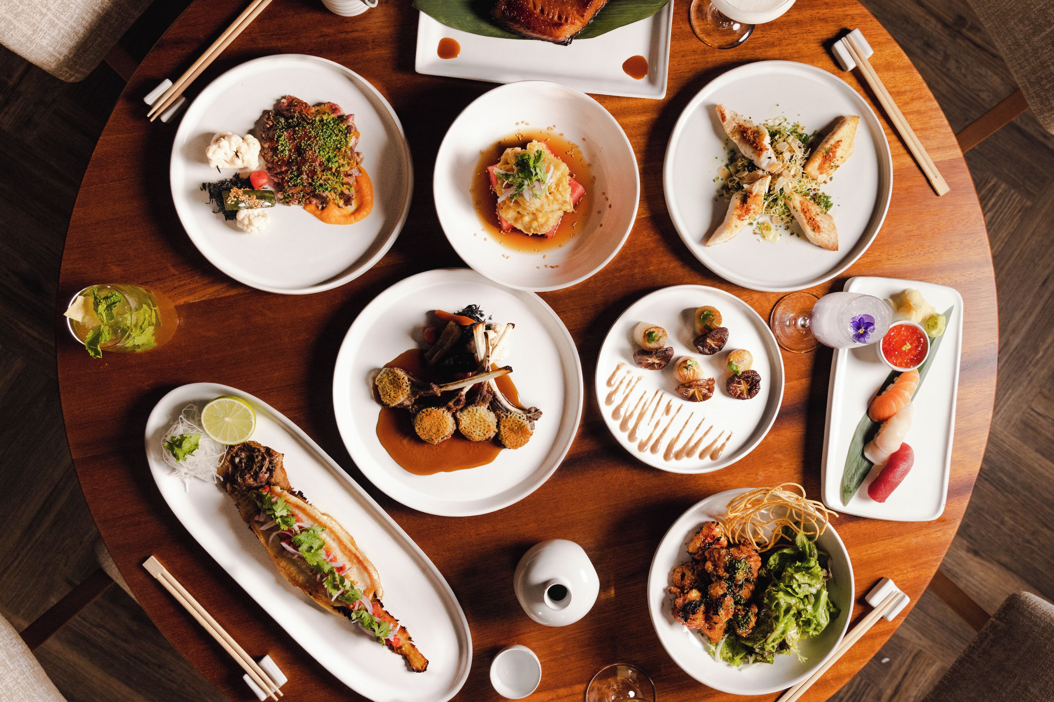 A variety of dishes on a wooden table, including seafood, vegetables, and meat, with chopsticks and a teapot visible.