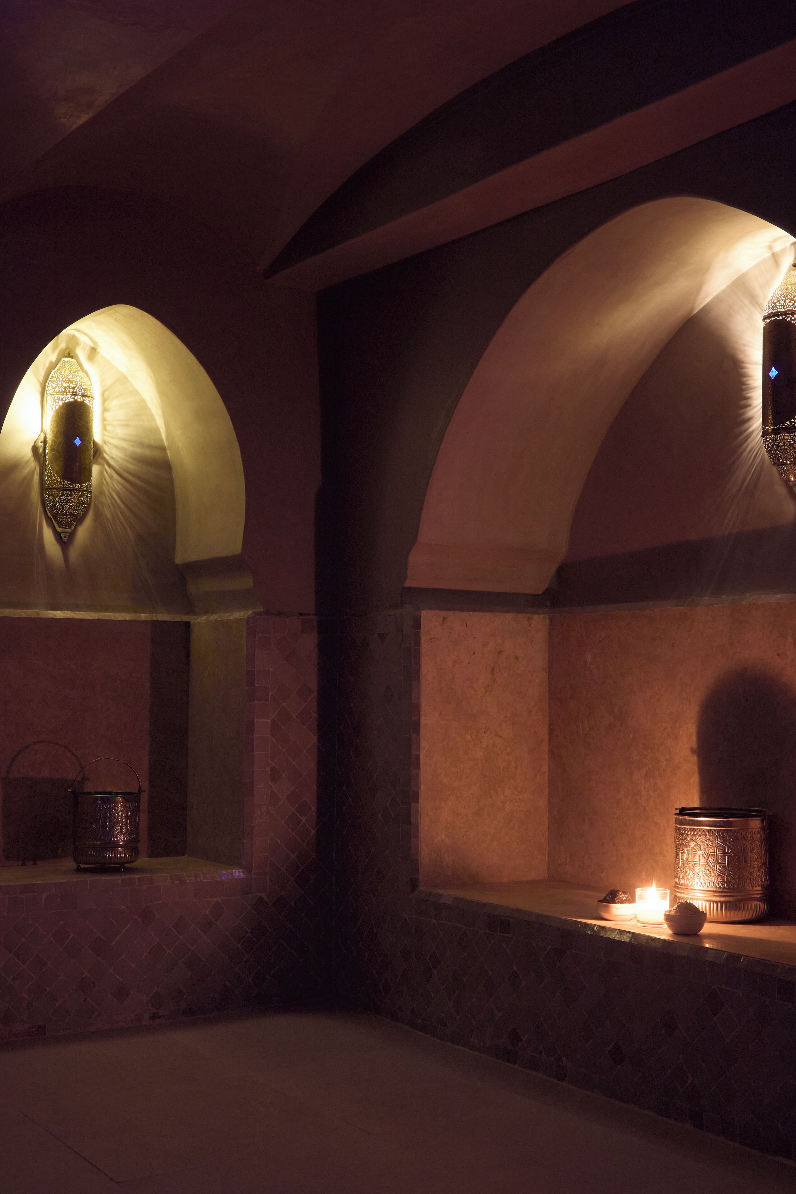 Un interior tenuemente iluminado, con arcos, suelo de baldosas y una vela sobre una estantería.