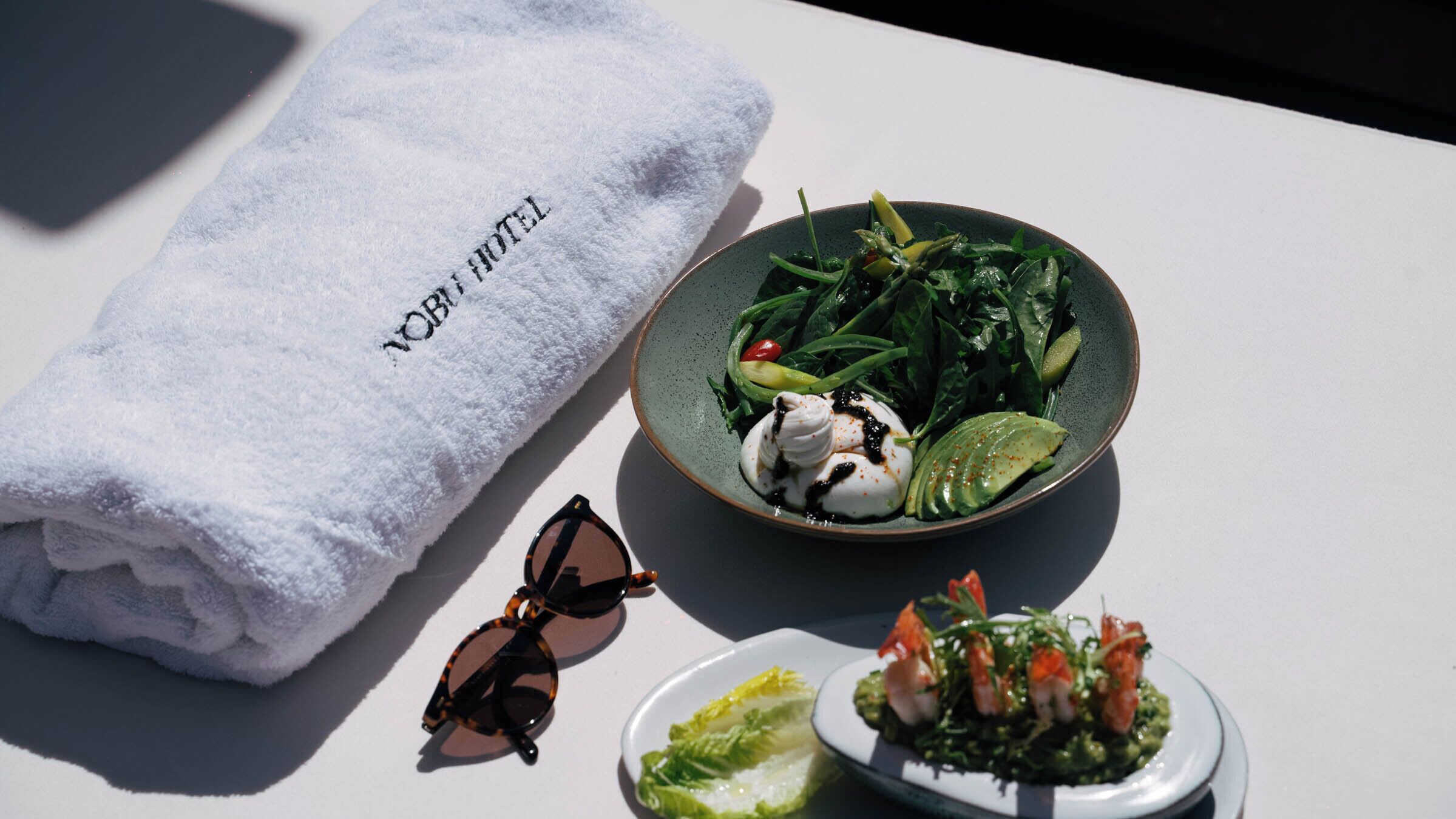 A neatly folded white towel with "HOTEL" embroidered on it, a bowl of salad with greens and a dollop of cream, and a small plate with a green sauce and shrimp garnish.