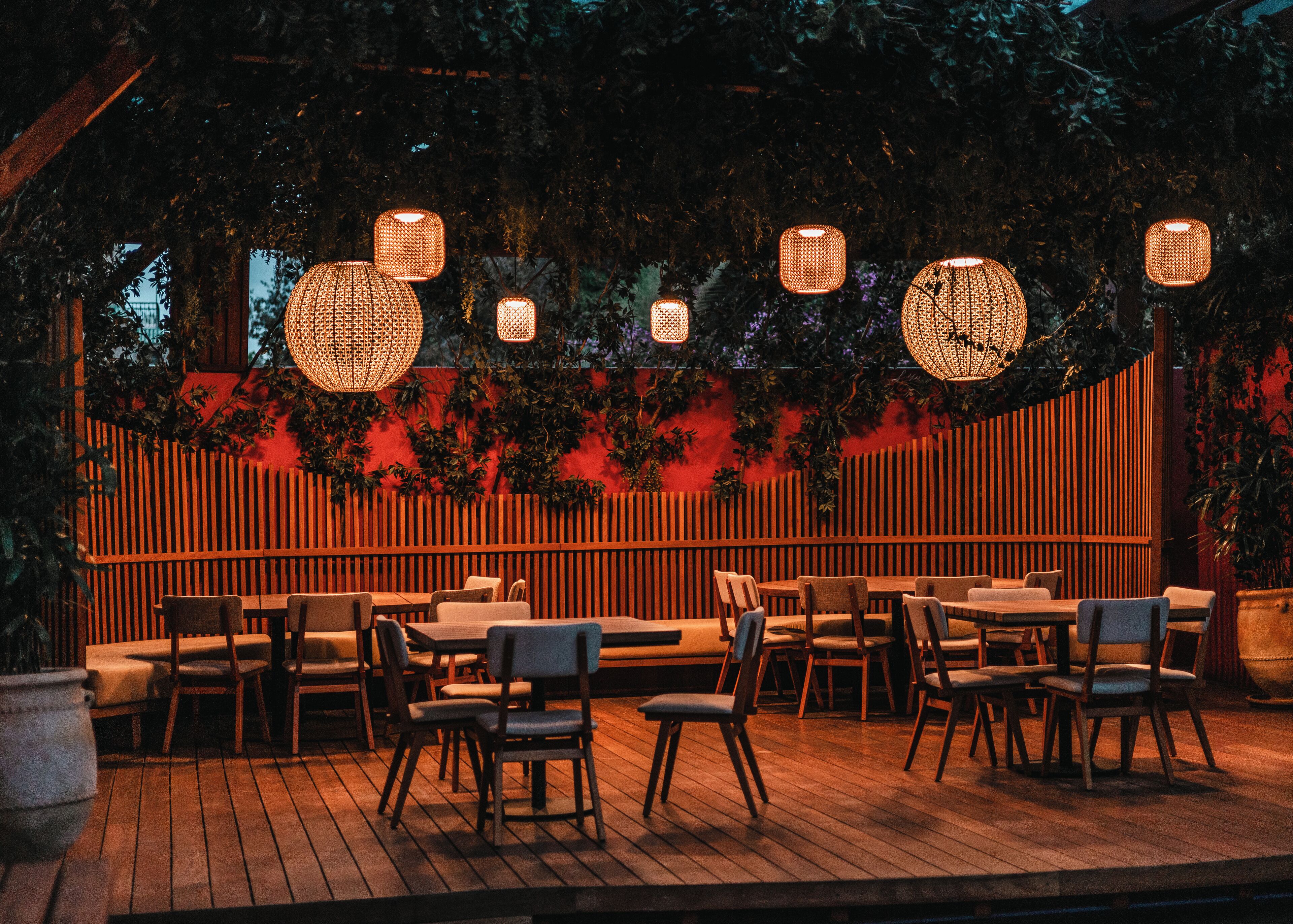 An outdoor dining area with wooden tables and chairs, surrounded by a wooden fence and foliage.