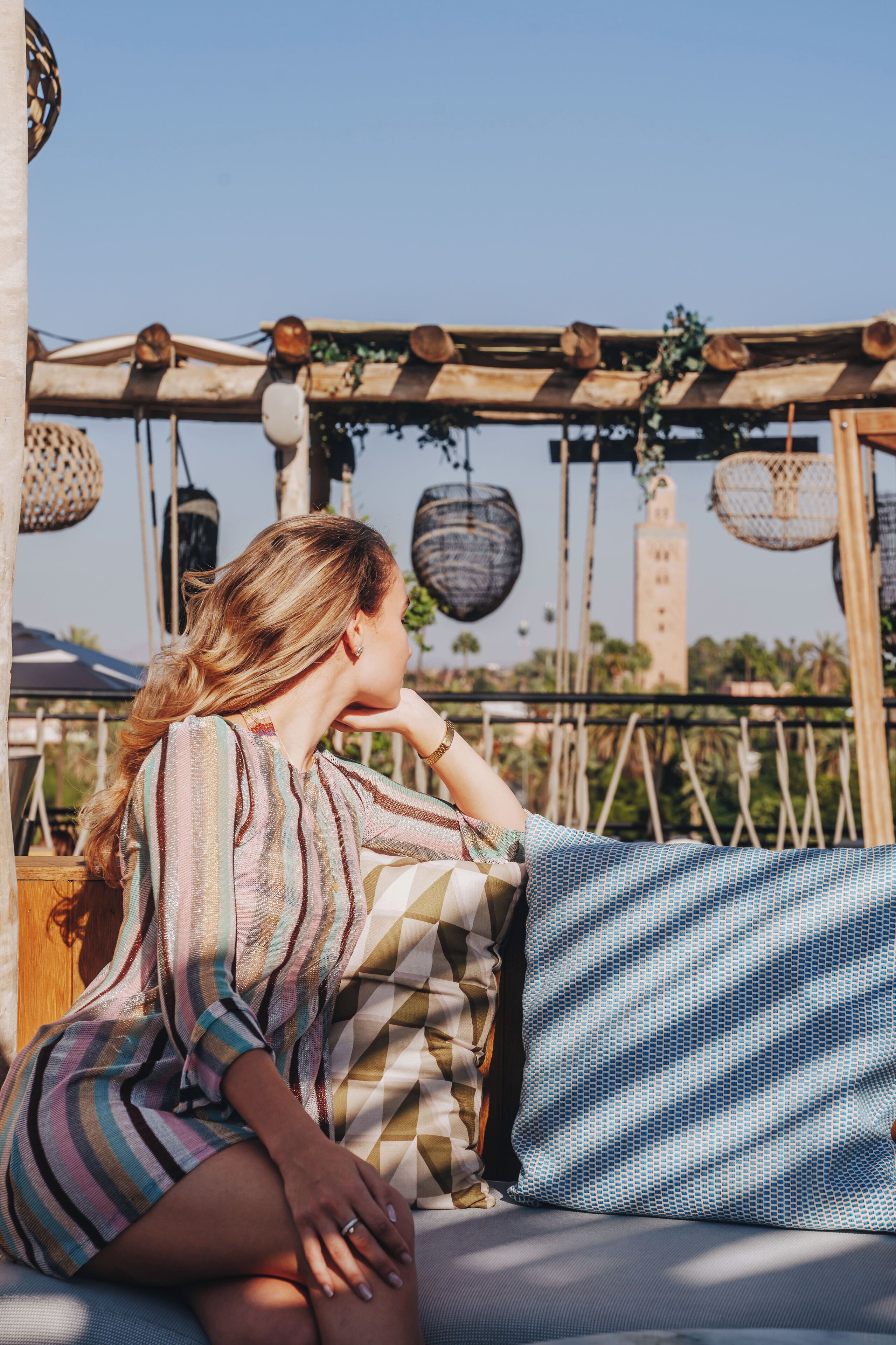 A person sitting on a bed with a patterned blanket, under a wooden structure with hanging baskets.