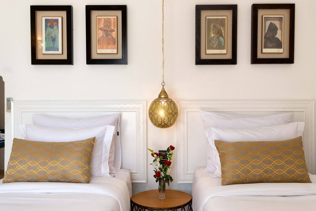Une chambre d’hôtel avec deux lits, une table ronde ornée d’un vase de fleurs et trois œuvres d’art encadrées accrochées au mur.