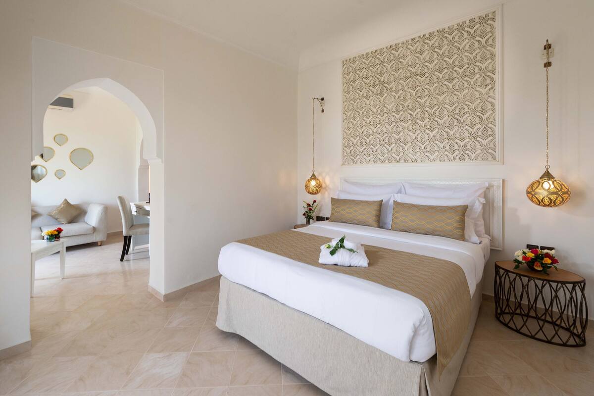 Une chambre d’hôtel moderne dotée d’un grand lit, d’un coin salon avec un canapé et un fauteuil, d’une décoration murale et d’une petite table d’appoint agrémentée de fleurs.