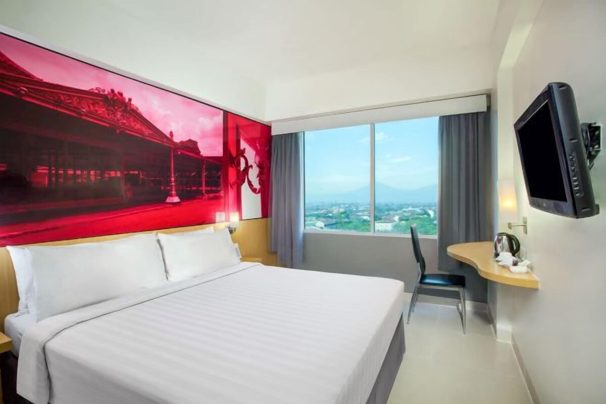 Habitación de hotel con una cama grande, un escritorio con silla, televisión y un ventanal con vistas a la ciudad.