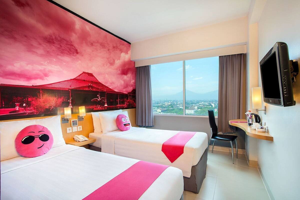 A hotel room with two beds, a desk, a TV, and a large window with a city view.