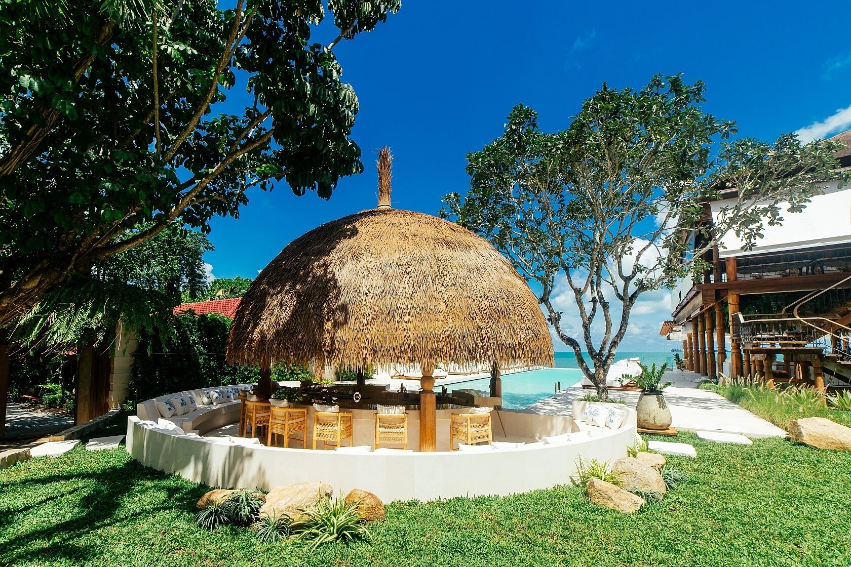 A thatched-roof gazebo with a circular seating area, surrounded by lush greenery and a view of the ocean.
