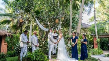 A wedding ceremony in a lush garden with a thatched-roof structure and white flowers.