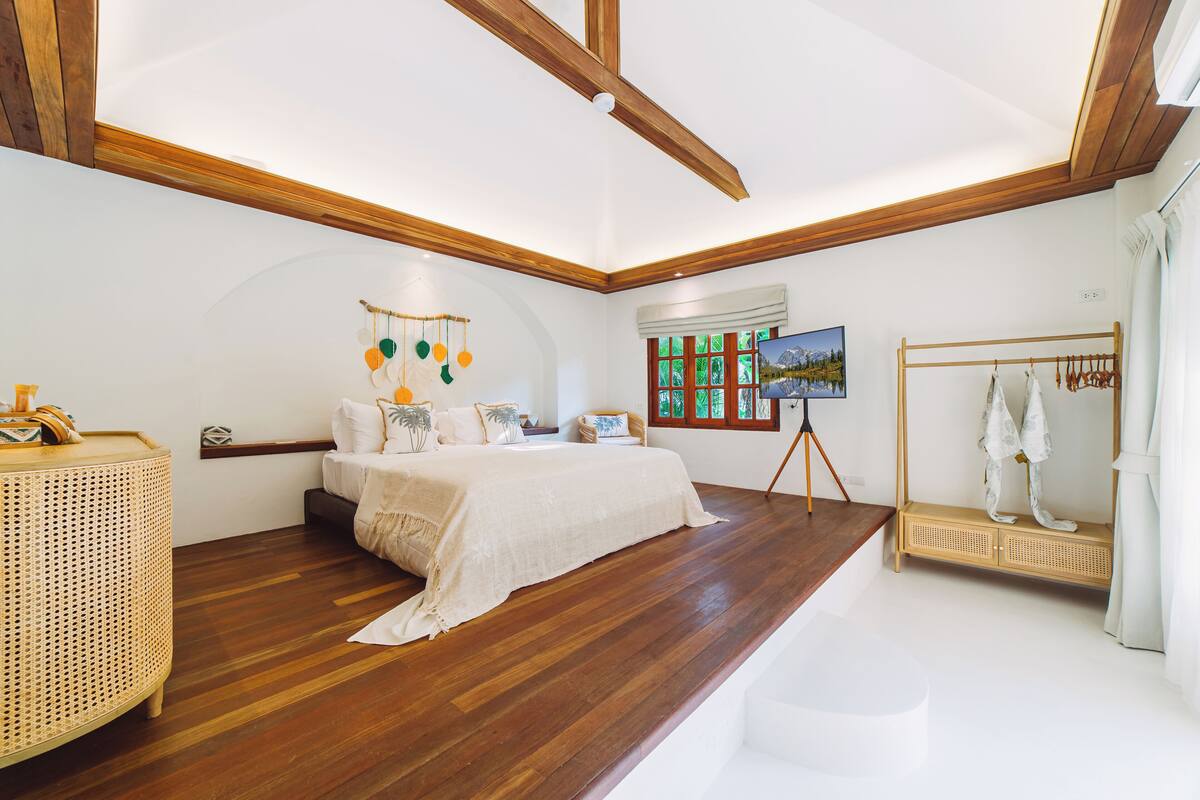 A bedroom with a bed, wooden floor, a rattan side table, a painting on the wall, and a clothing rack.
