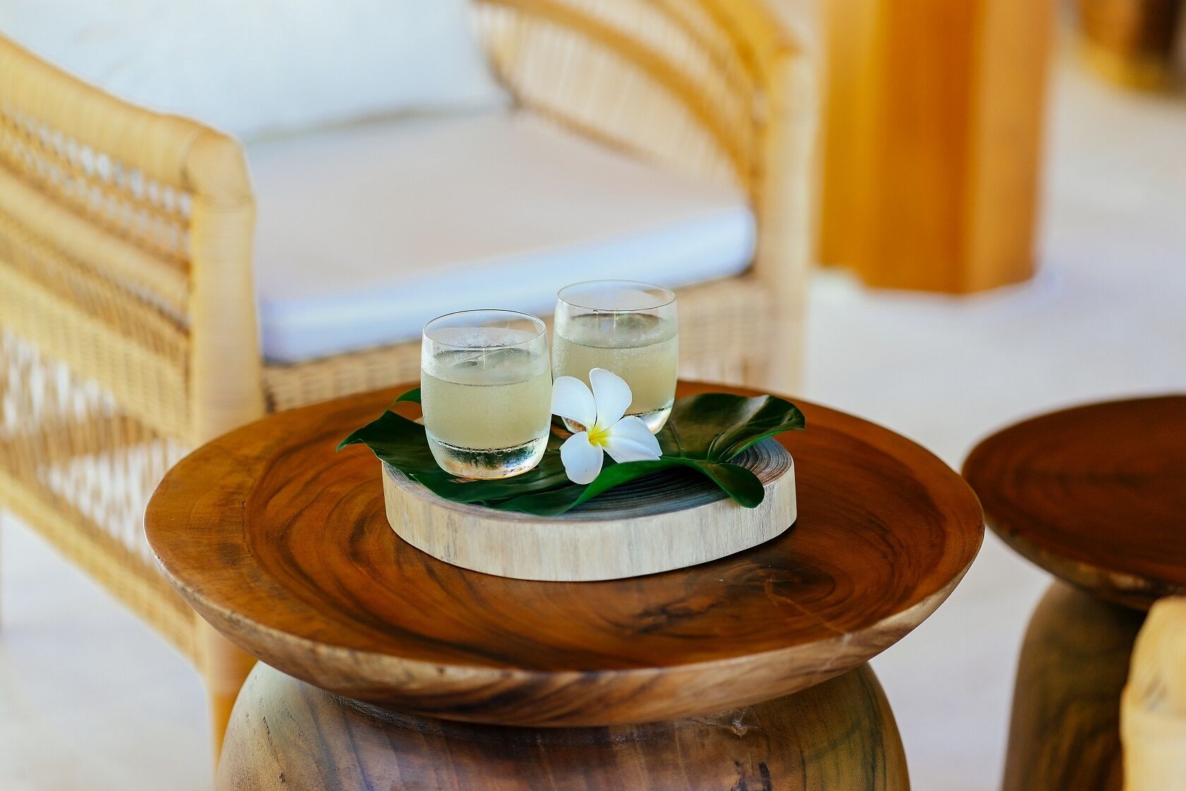 A wooden table with two glasses of a light-colored beverage, a white flower, and a green leaf on a wooden coaster.