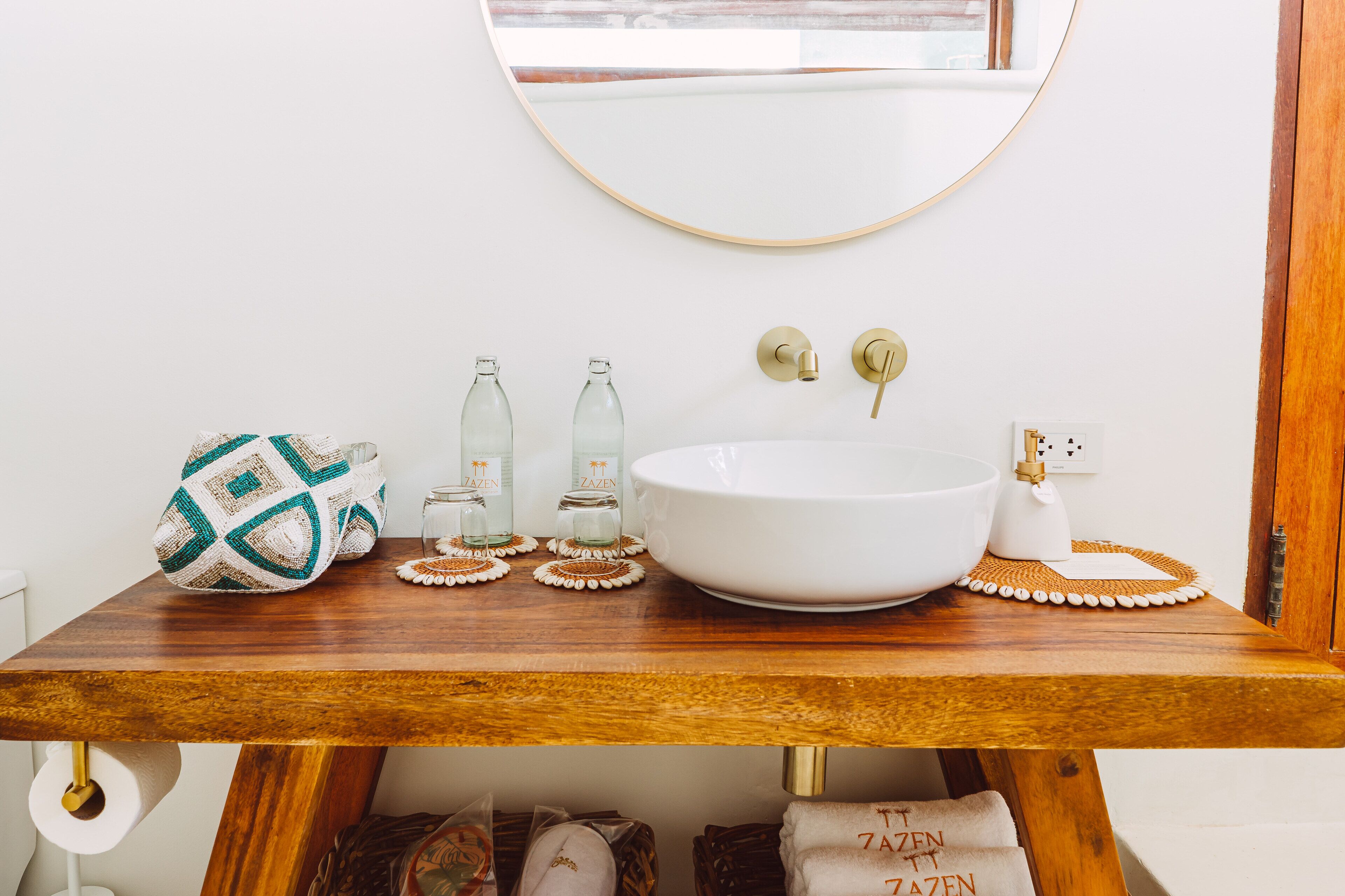 Une salle de bain avec un meuble-lavabo en bois, un grand lavabo blanc et un miroir au-dessus. On y trouve deux bouteilles en verre et un bol décoratif sur le meuble-lavabo.