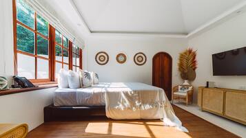 Ampia camera da letto con un letto grande, pavimento in legno, finestra con persiane, televisore e tavolino in rattan.