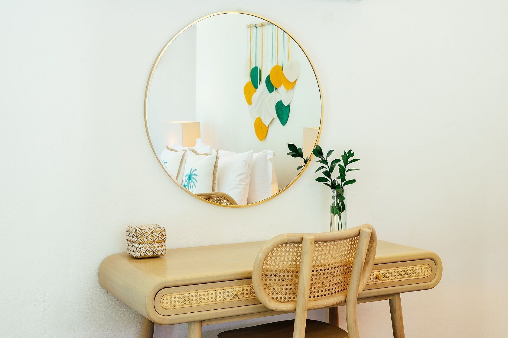 A room with a round mirror, a rattan chair, a small table with a decorative item, and a plant in a clear vase.