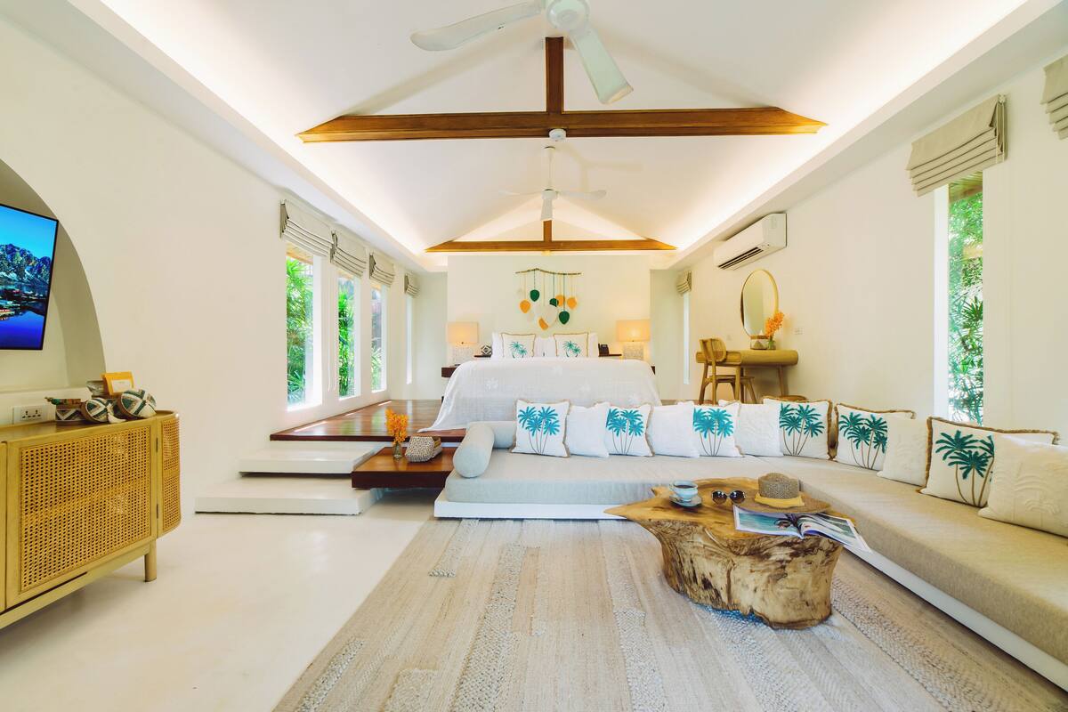 A spacious bedroom with a large bed, a wooden ceiling fan, and a view of greenery outside.