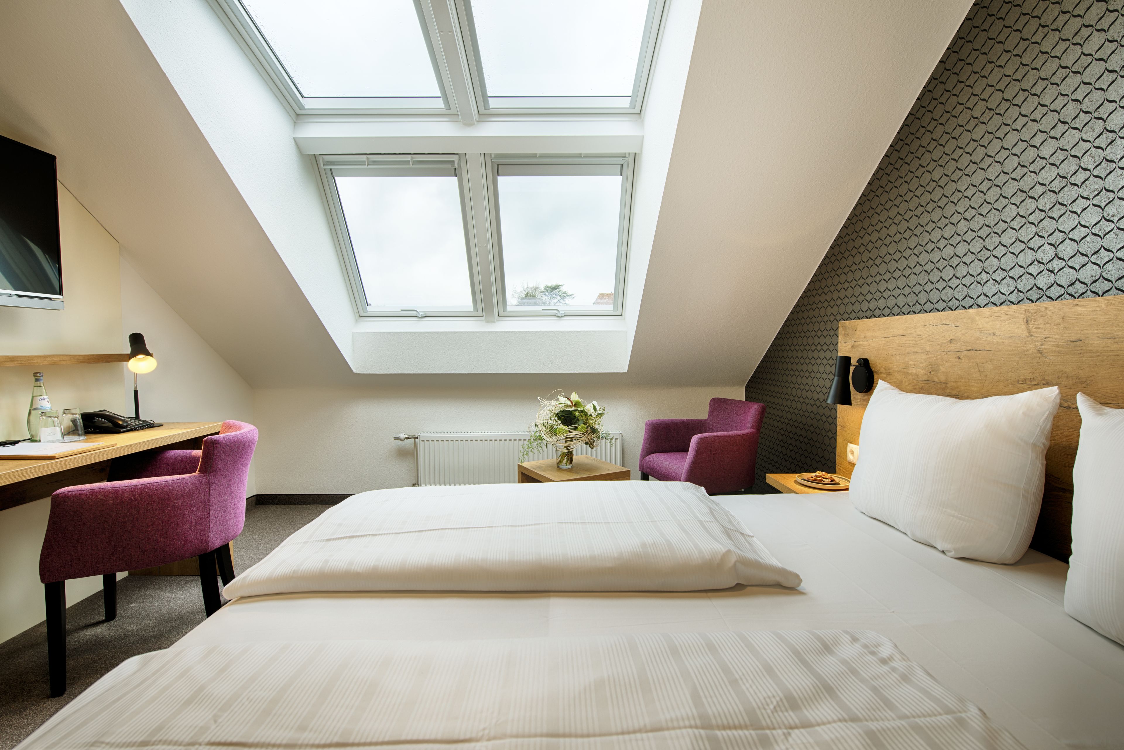 Een slaapkamer met dakraam, een bed, bureau, stoel en een klein tafeltje met een bloevas.