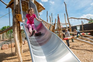 A child sliding down a metal slide at a playground with wooden structures and ropes. - Das Prinzregent (Edenkoben)