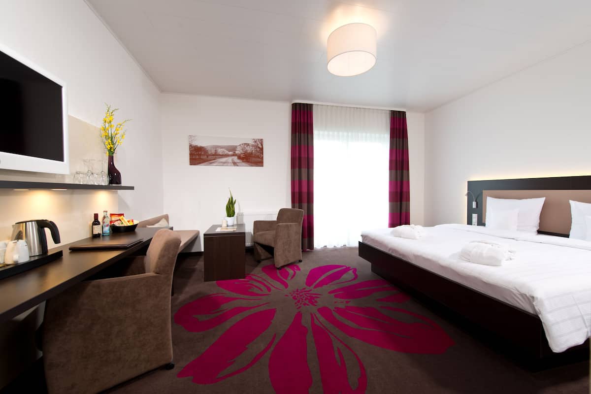 A hotel room with a bed, a desk with a flat-screen TV, a chair, a small table with a plant, and a large floral carpet.