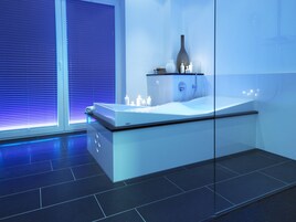 A modern bathroom with a glass-enclosed bathtub, white fixtures, and a large window with blinds.