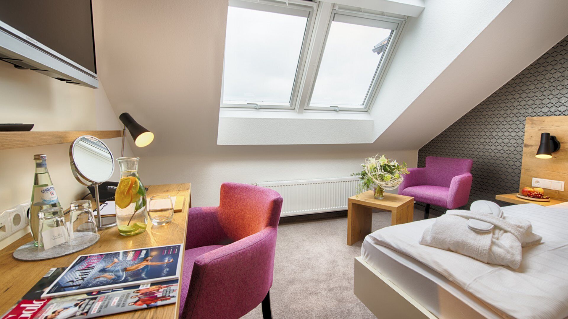 Une chambre d’hôtel moderne, équipée d’un lit, d’un bureau avec un miroir, d’une chaise et d’une petite table sur laquelle est posé un vase de fleurs.
