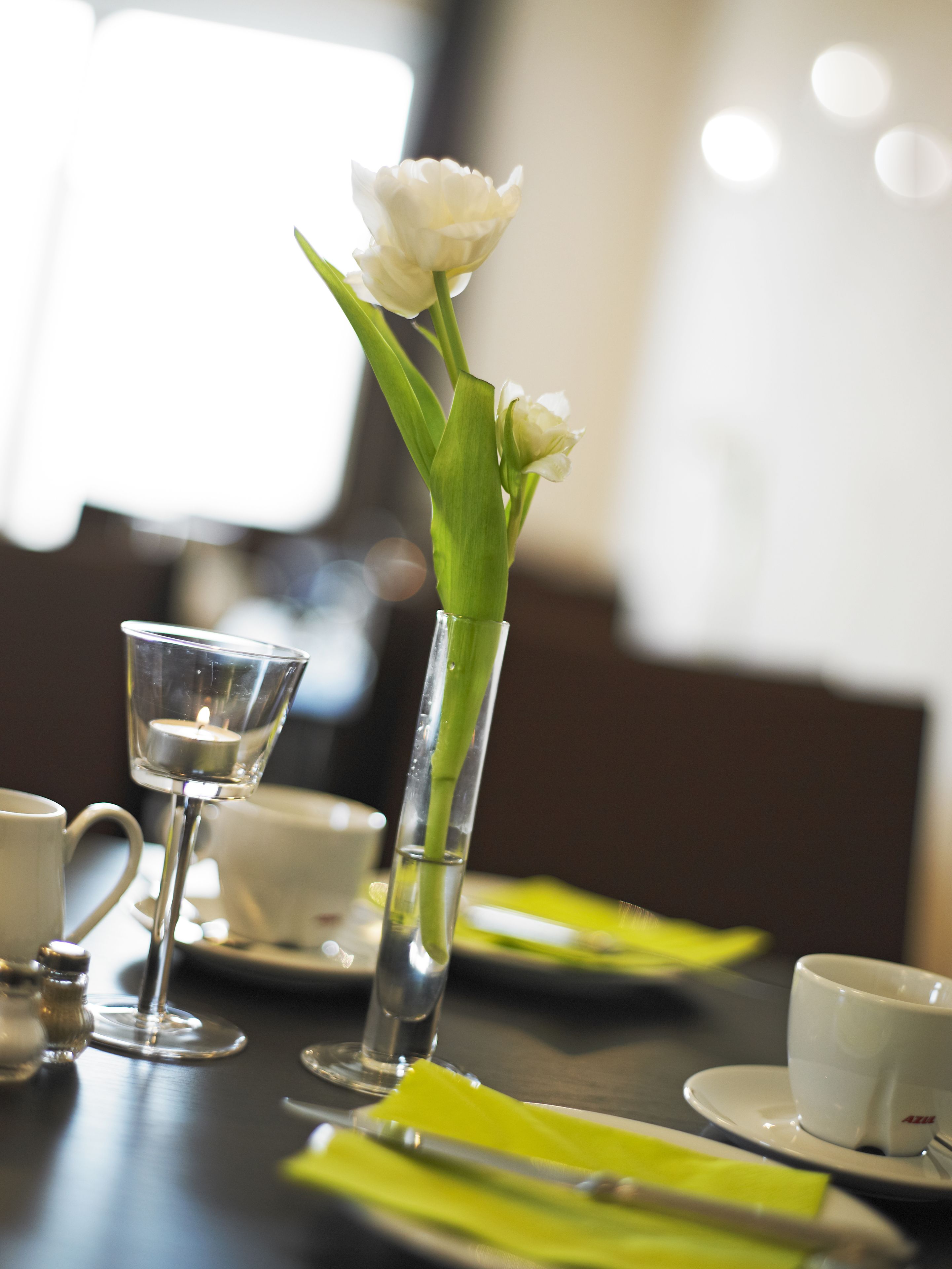 A dining table with a candle in a glass holder, a white flower in a clear vase, and a white cup and saucer.