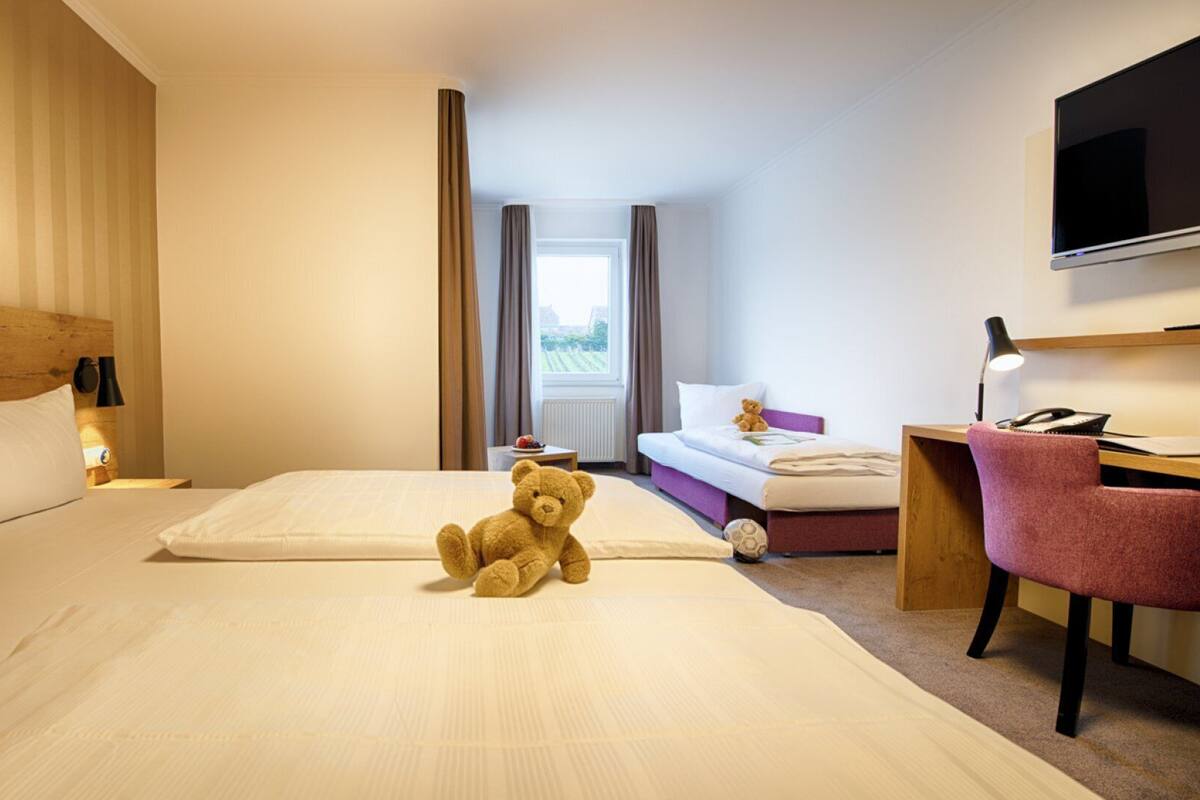 A hotel room with a large bed, a desk with a chair, a television, and a teddy bear on the bed.