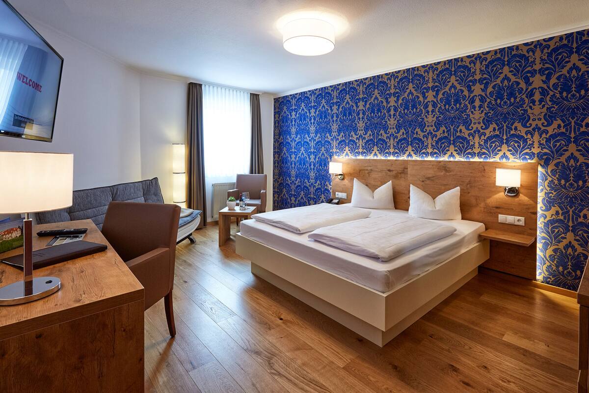 A hotel room with a large bed, a desk with a lamp, a chair, a TV, and a wooden headboard.