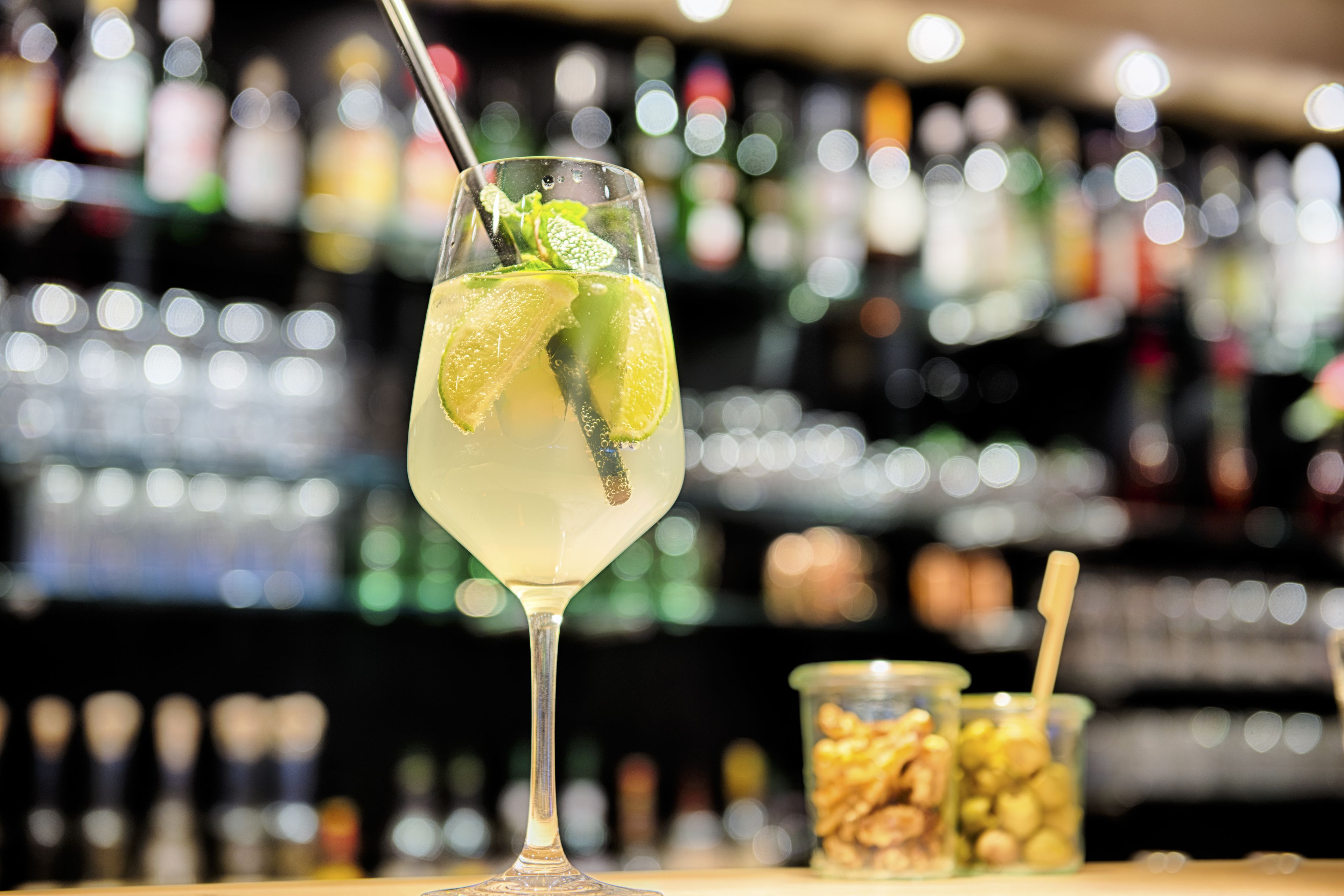 A cocktail glass with lime and mint garnish, a straw, and a spoon, with a blurred background of bottles.