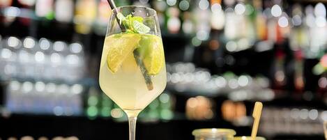 A cocktail glass with lime and mint garnish, a straw, and a spoon, with a blurred background of bottles.