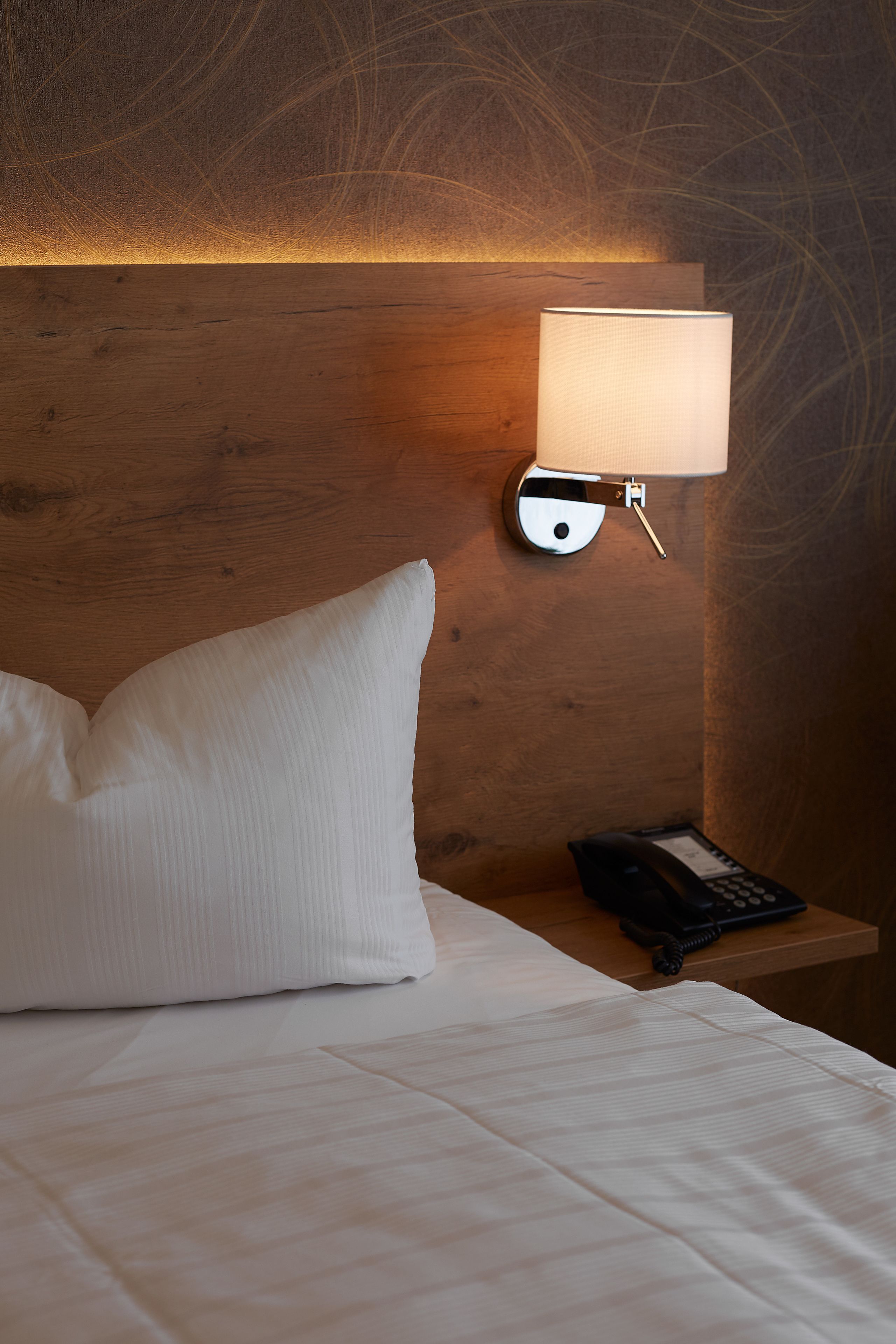 A neatly made bed with white linens and a single white pillow. A wall-mounted lamp with a white shade is visible on the wooden headboard.