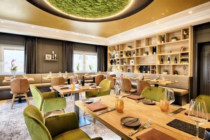 A modern restaurant with wooden tables and green chairs, a wall of wine bottles, and a green textured ceiling.
