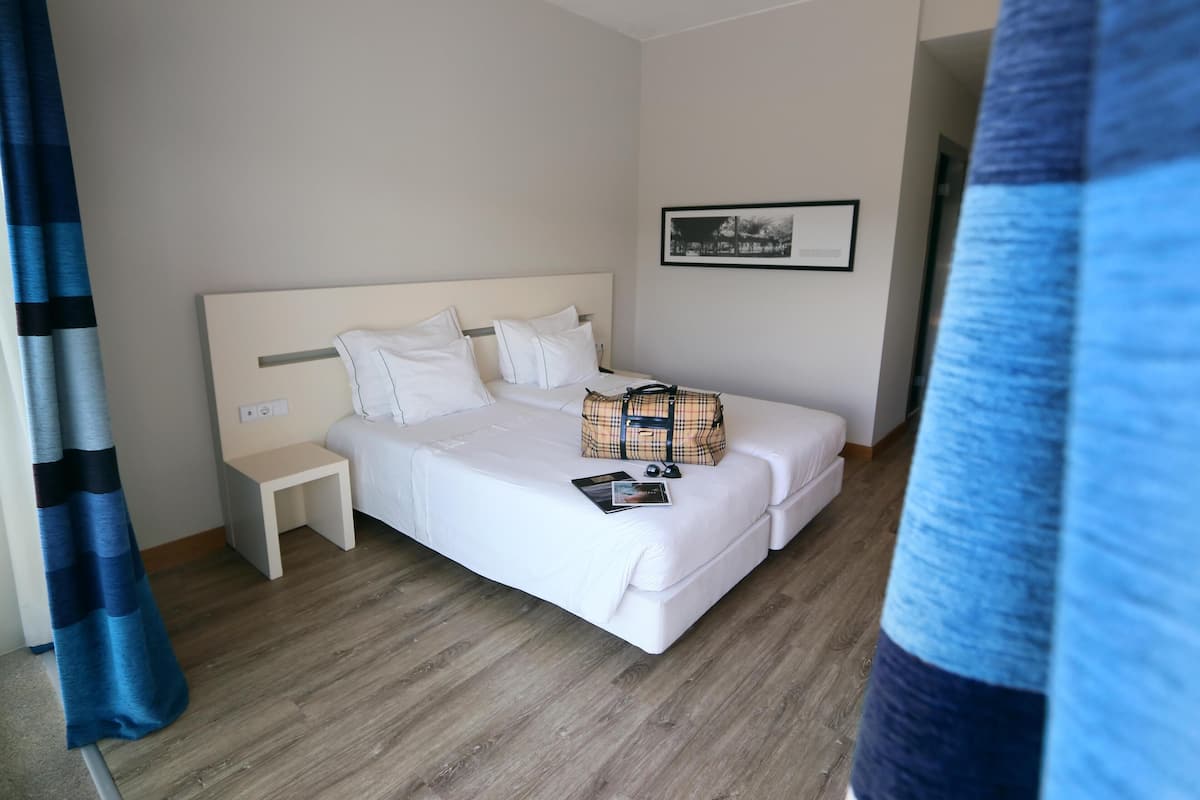 Ett hotellrum med en säng, ett sängbord, ett litet sidobord, en rutig väska och en inramad tavla på väggen.
