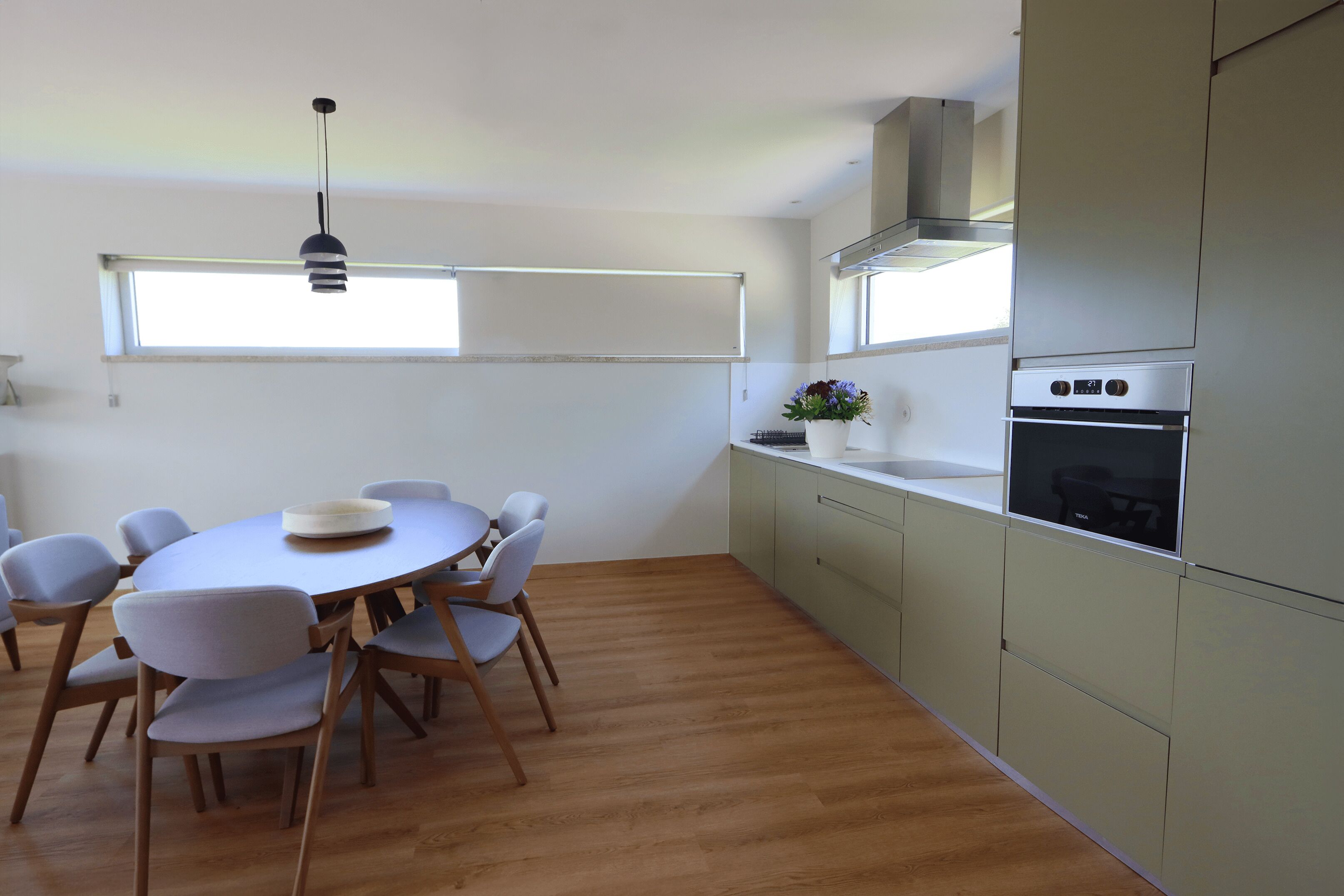 A modern kitchen with a round dining table, light-colored chairs, a built-in oven, and a window above the sink.