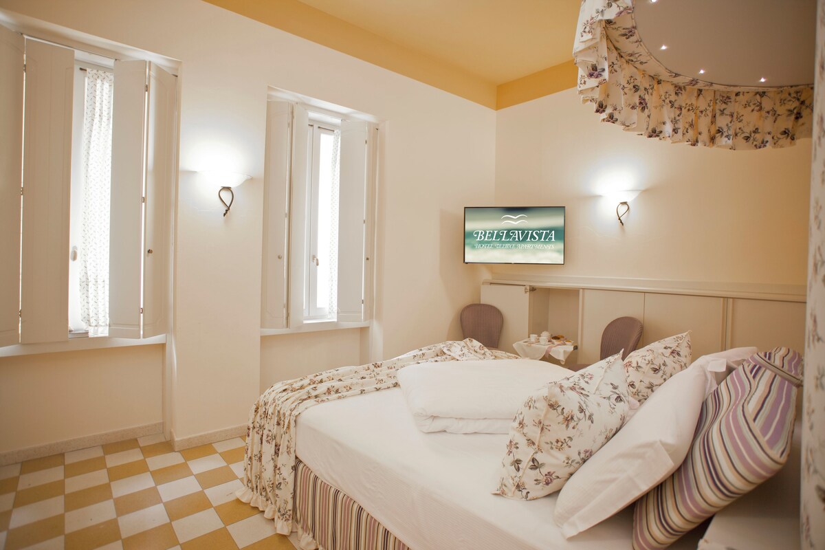 Een hotelkamer met een zwart-wit schakeerd motief vloer, een bed met bloemenprint beddengoed, een televisie aan de muur en een raam met gordijnen.
